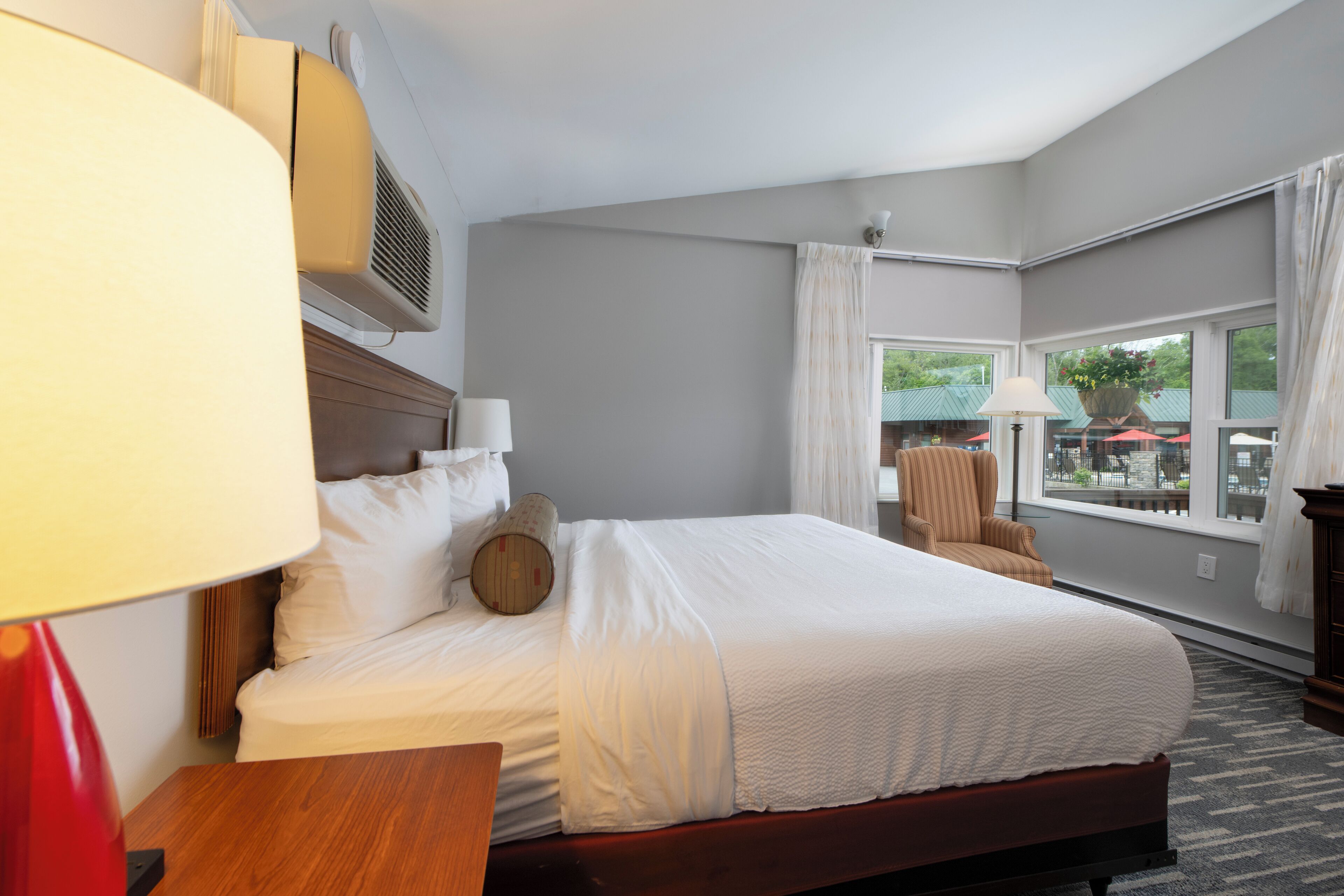A hotel room with a large bed, a wooden nightstand, a red lamp, a window with curtains, and a view of the outside.