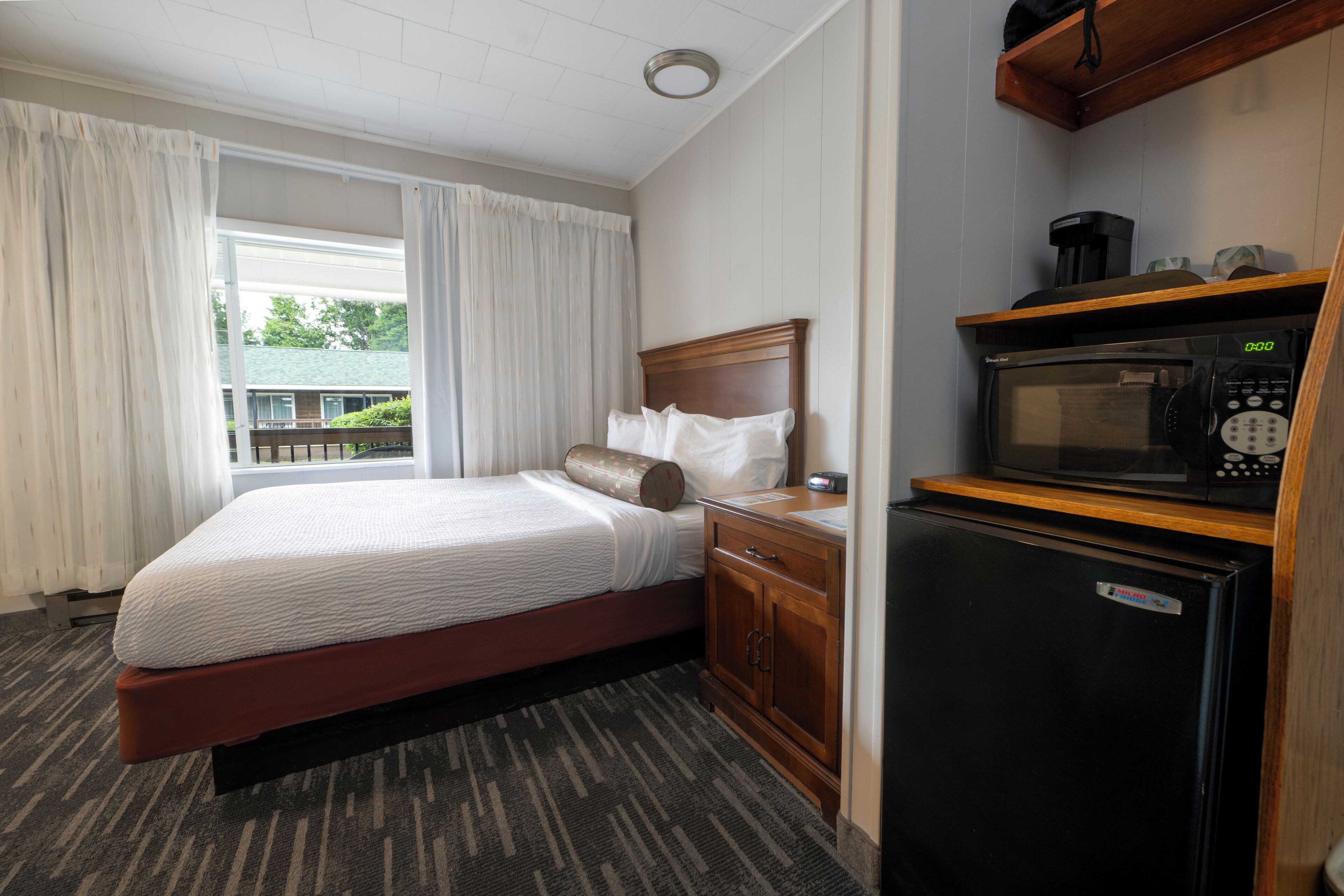 A hotel room with a bed, a small kitchenette with a microwave, and a window with curtains.