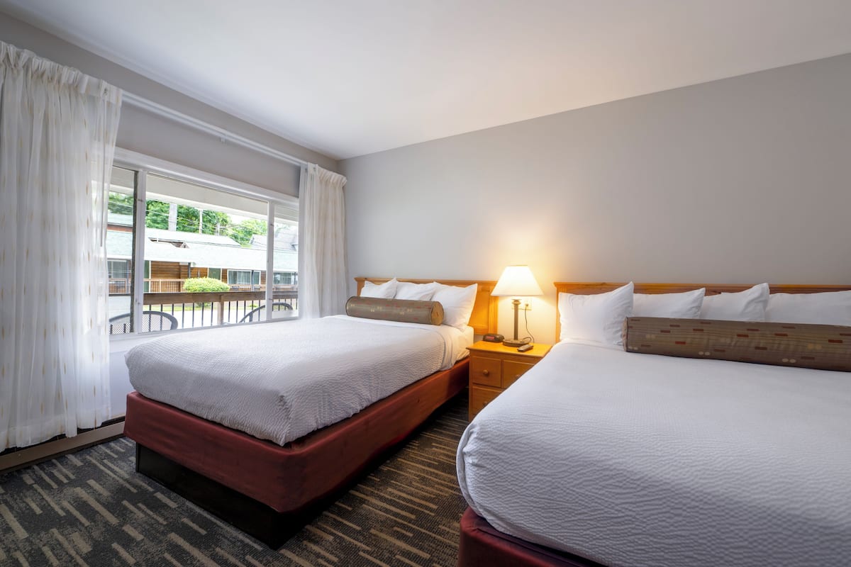 A hotel room with two beds, a window with curtains, a nightstand with a lamp, and a view of the outside balcony.