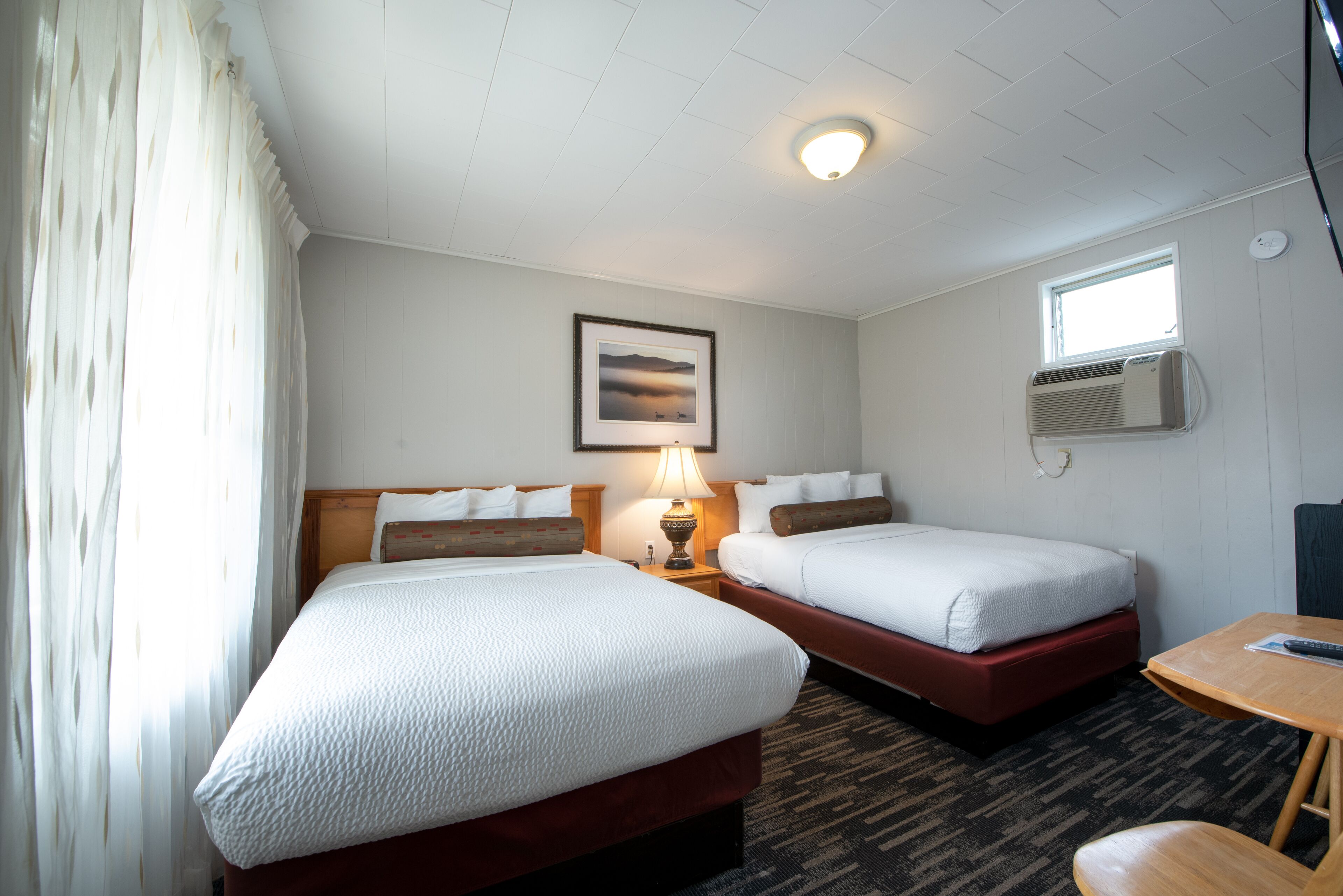 A hotel room with two beds, a window, an air conditioning unit, a lamp, and a small table.