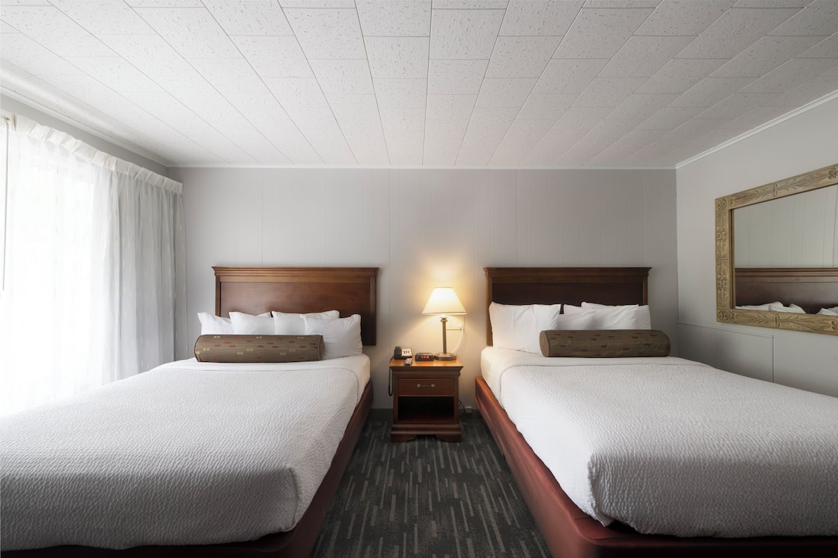 A hotel room with two beds, a nightstand with a lamp, and a large mirror on the wall.