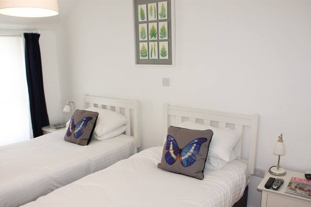 A hotel room with two single beds, a nightstand with a lamp, a small table with a magazine, and a window with a curtain.