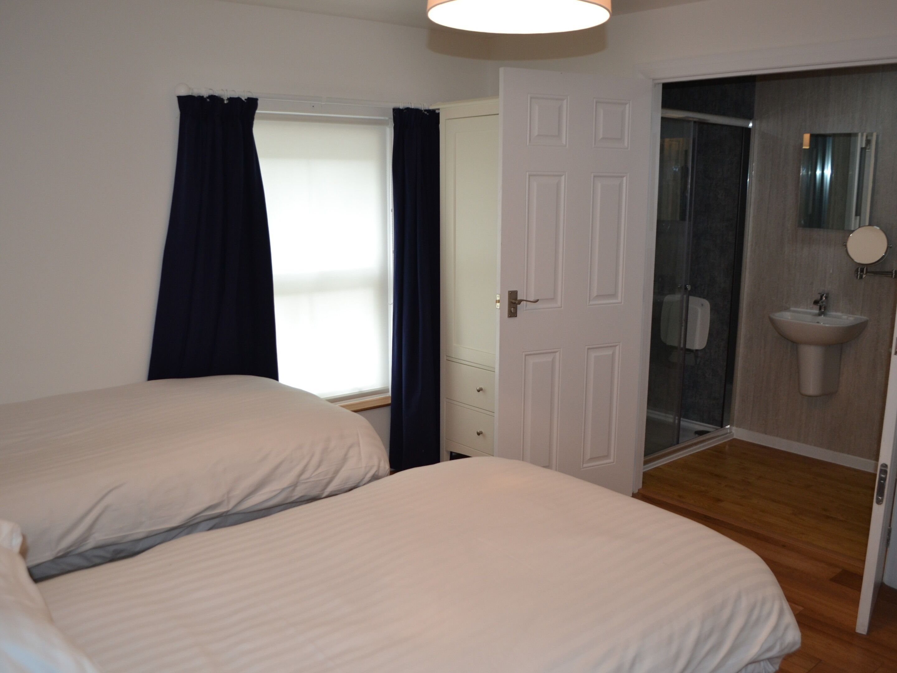 Un dormitorio con dos camas, una ventana con cortinas, una puerta que da al baño y una zona de ducha.