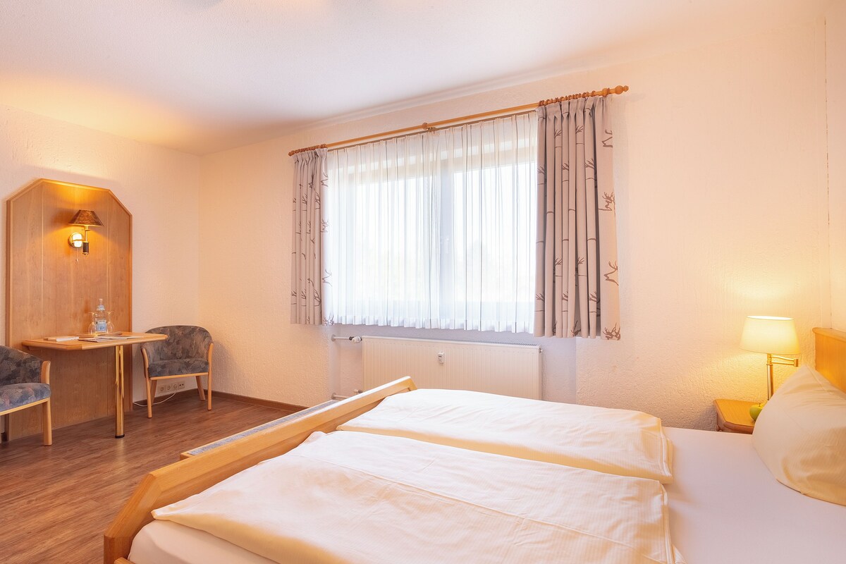 Habitación de hotel con cama, escritorio, silla, ventana con cortinas y lámpara de pared.