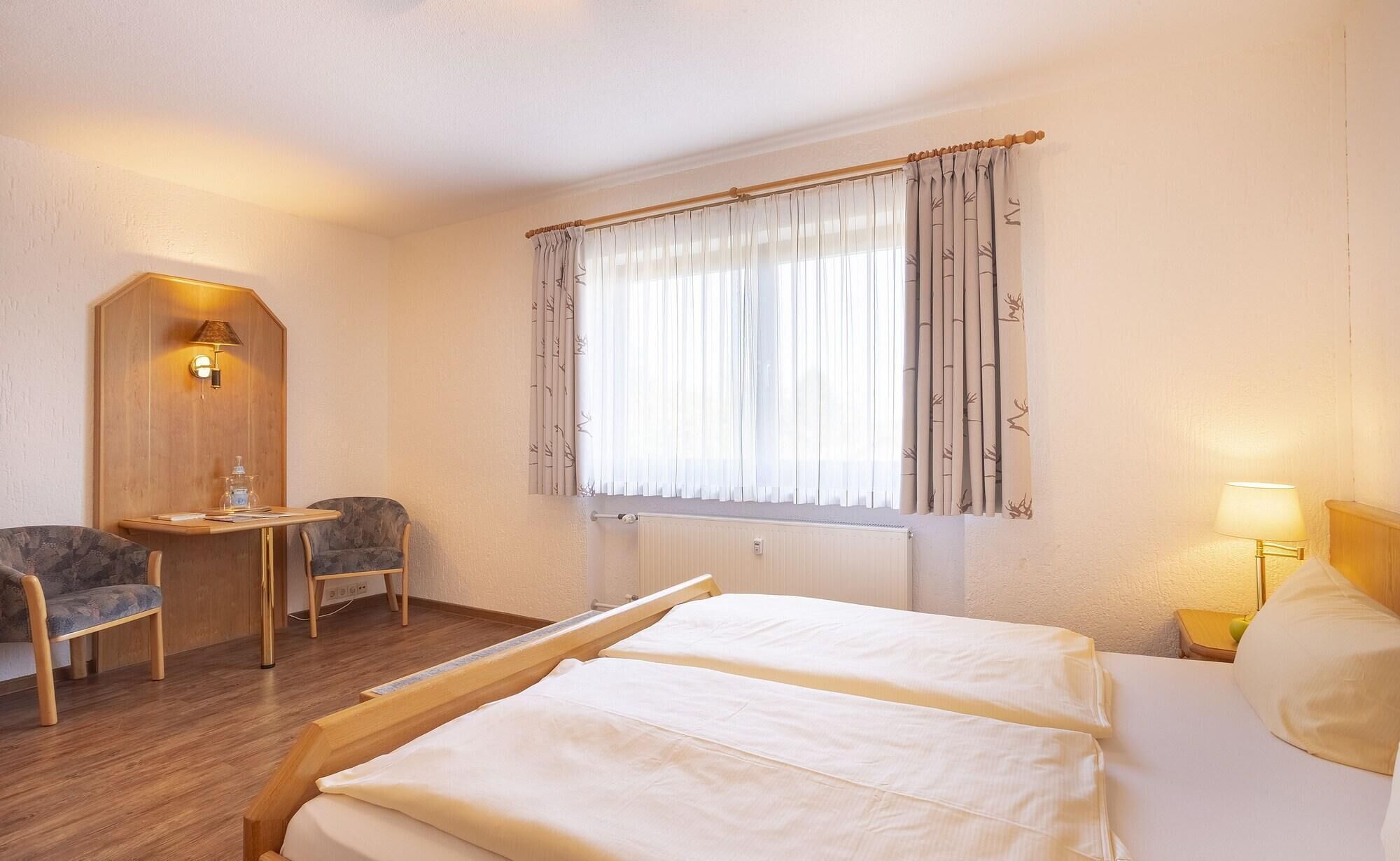 A hotel room with a bed, a desk, a chair, a window with curtains, and a wall-mounted lamp.
