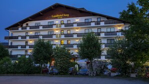 A multi-story hotel with a sign reading "Landhotel Seeg". It features balconies with flower boxes, multiple windows, and a parking area in front.