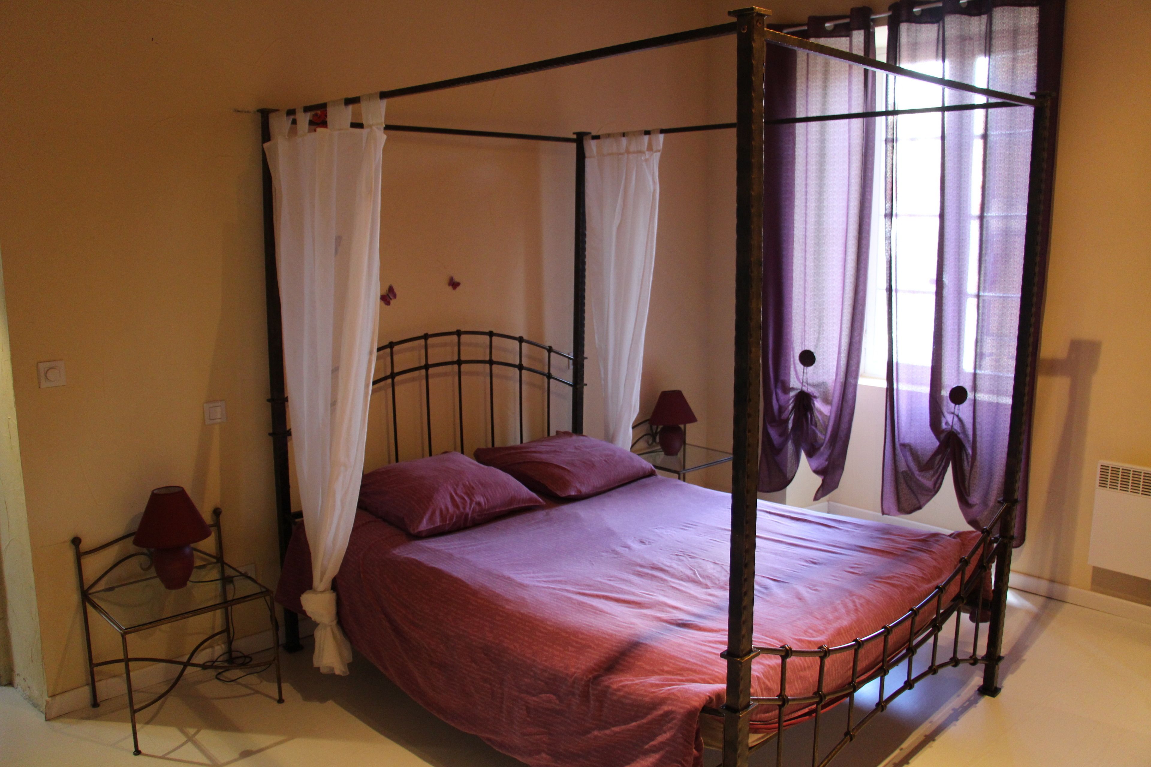 Una cama con dosel y cuatro columnas, una mesita de noche con lámpara, y un calefactor fijado a la pared.