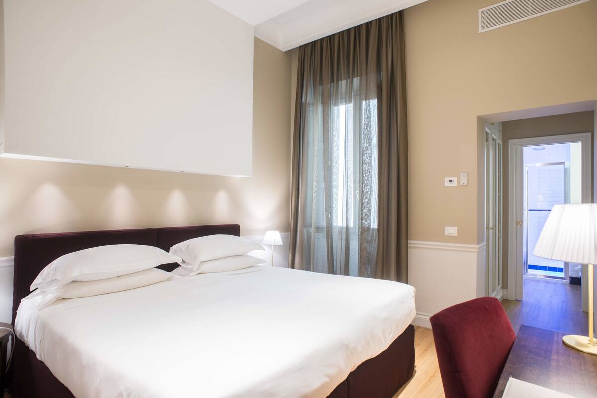 A hotel room with a bed, a chair, a lamp, a window with curtains, and a door leading to another room.