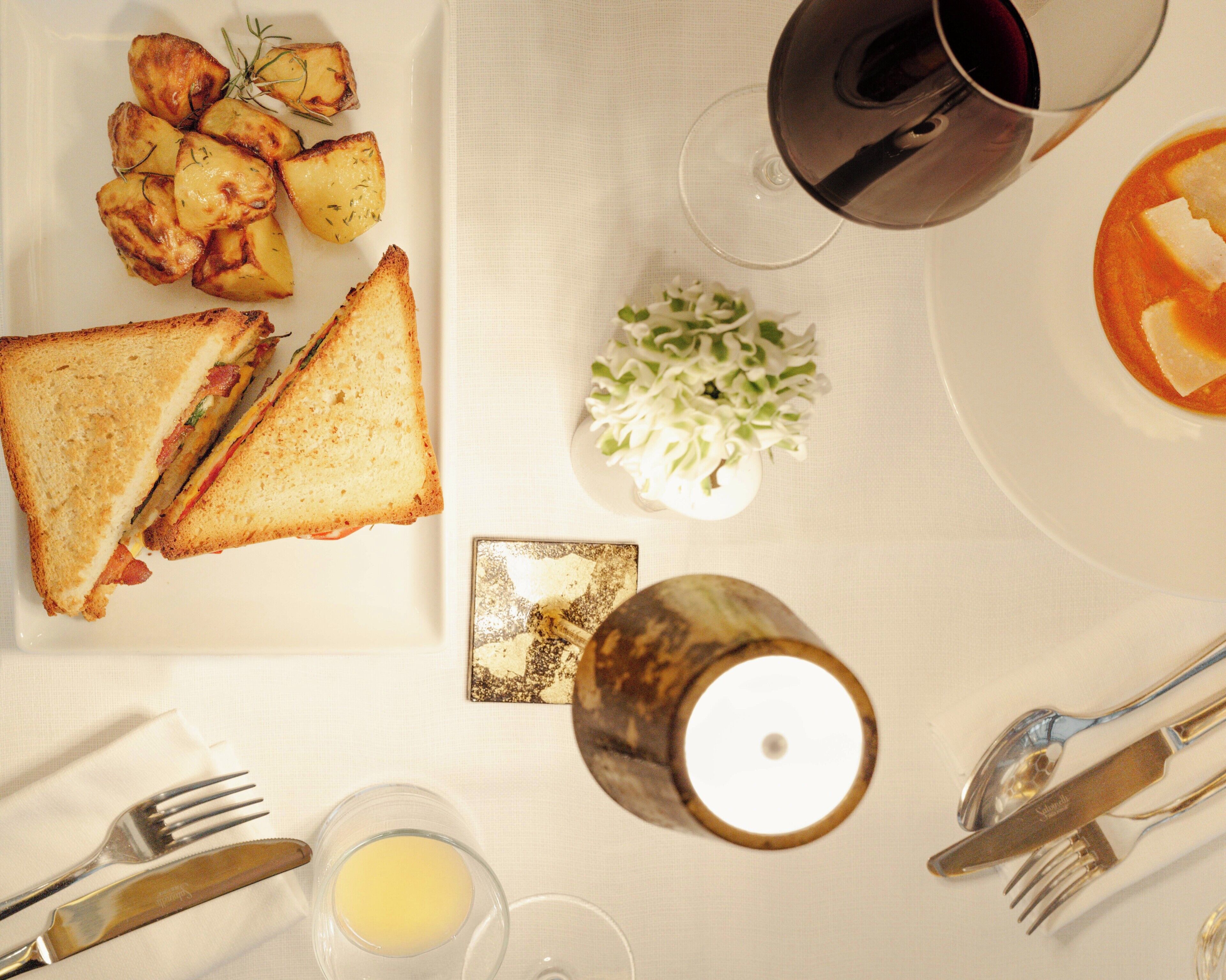 Mesa posta com sanduíche, batatas assadas, salada pequena, vela e copo de vinho tinto, além de talheres.