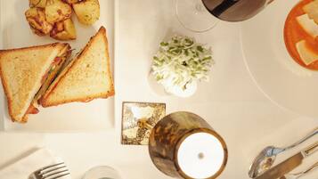 Mesa posta com sanduíche, batatas assadas, salada pequena, vela e copo de vinho tinto, além de talheres.