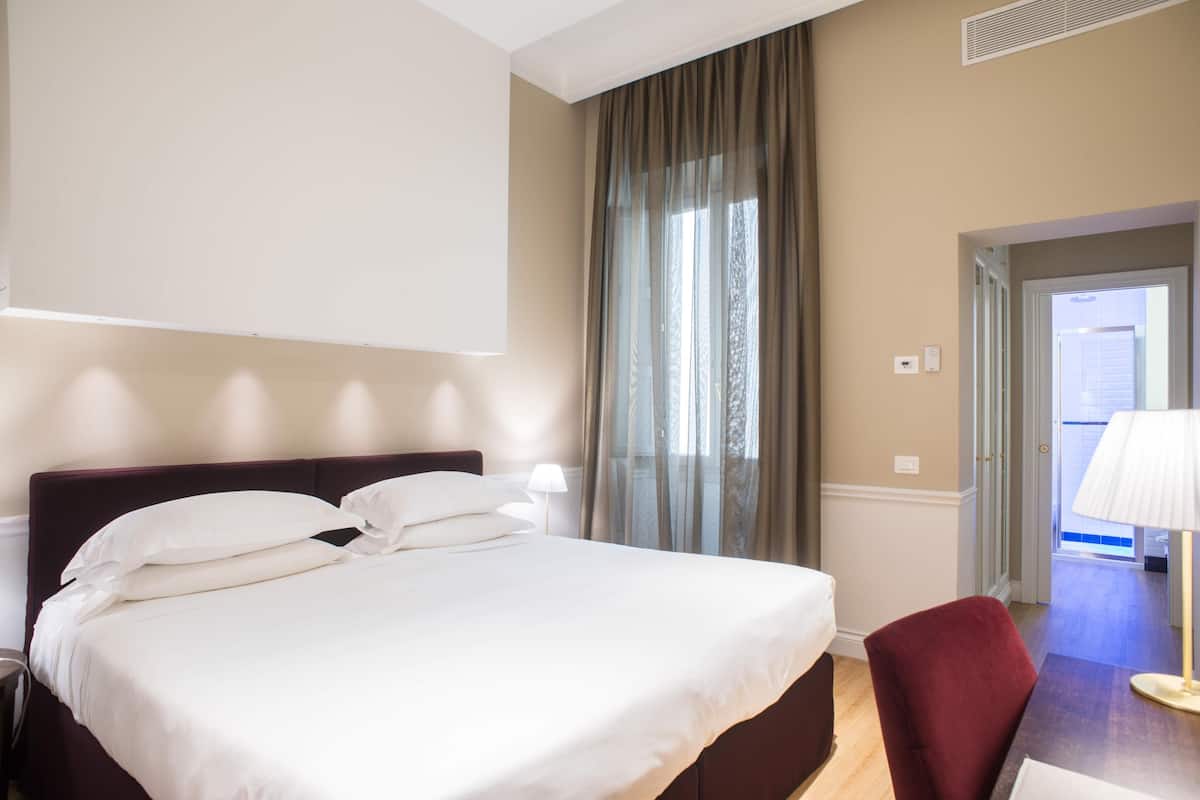 Une chambre d’hôtel avec un lit, une chaise, une lampe, une fenêtre avec des rideaux et une porte donnant sur une autre pièce.