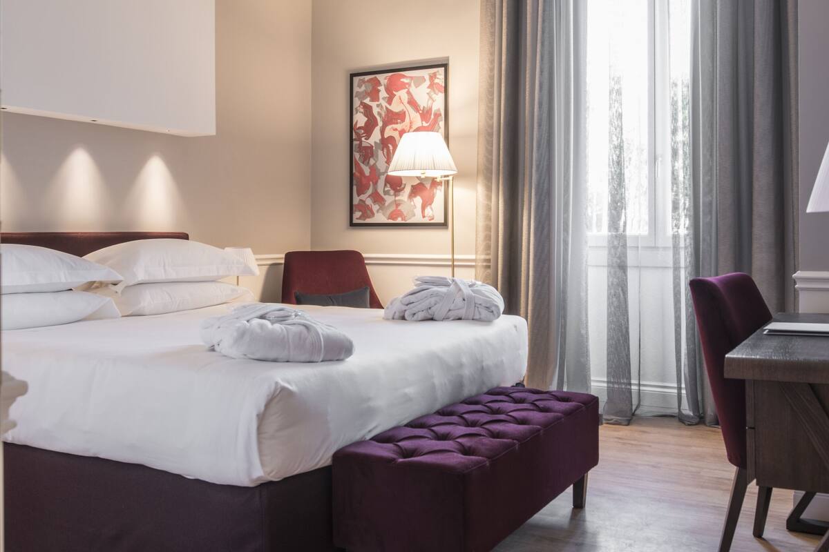 A hotel room with a bed, a desk, a chair, a window with curtains, and a painting on the wall.