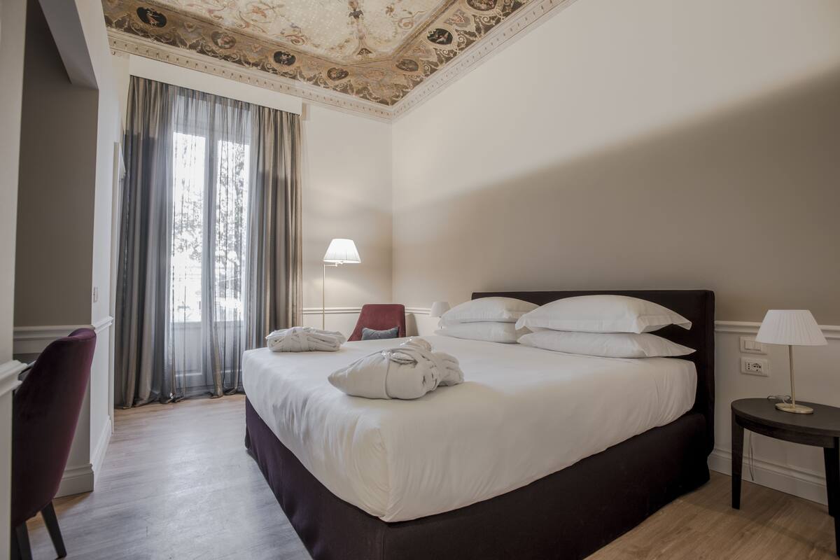 A hotel room with a large bed, a desk, a chair, a lamp, and a window with curtains.