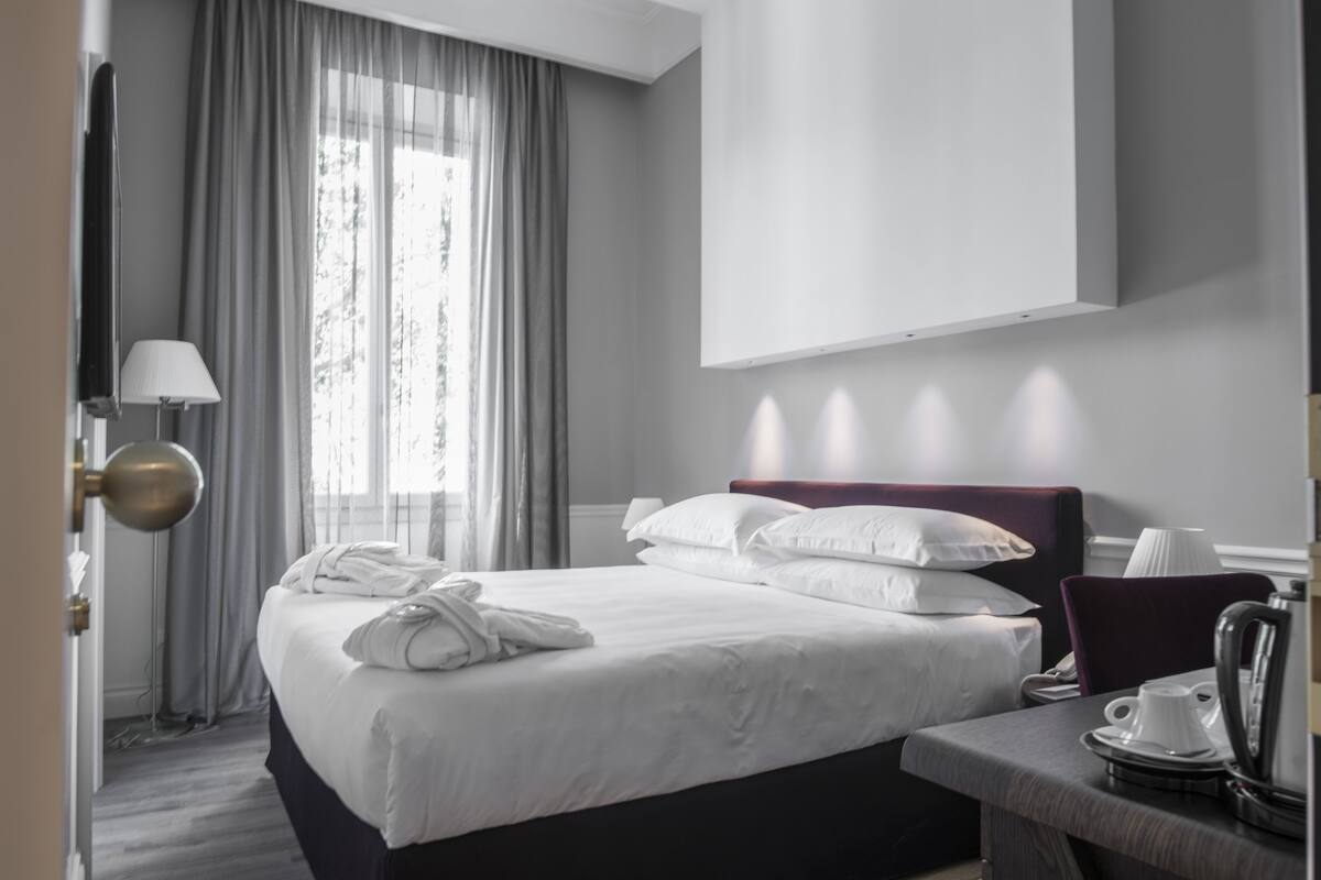 A hotel room with a bed, a desk with a kettle, a lamp, and a window with curtains.