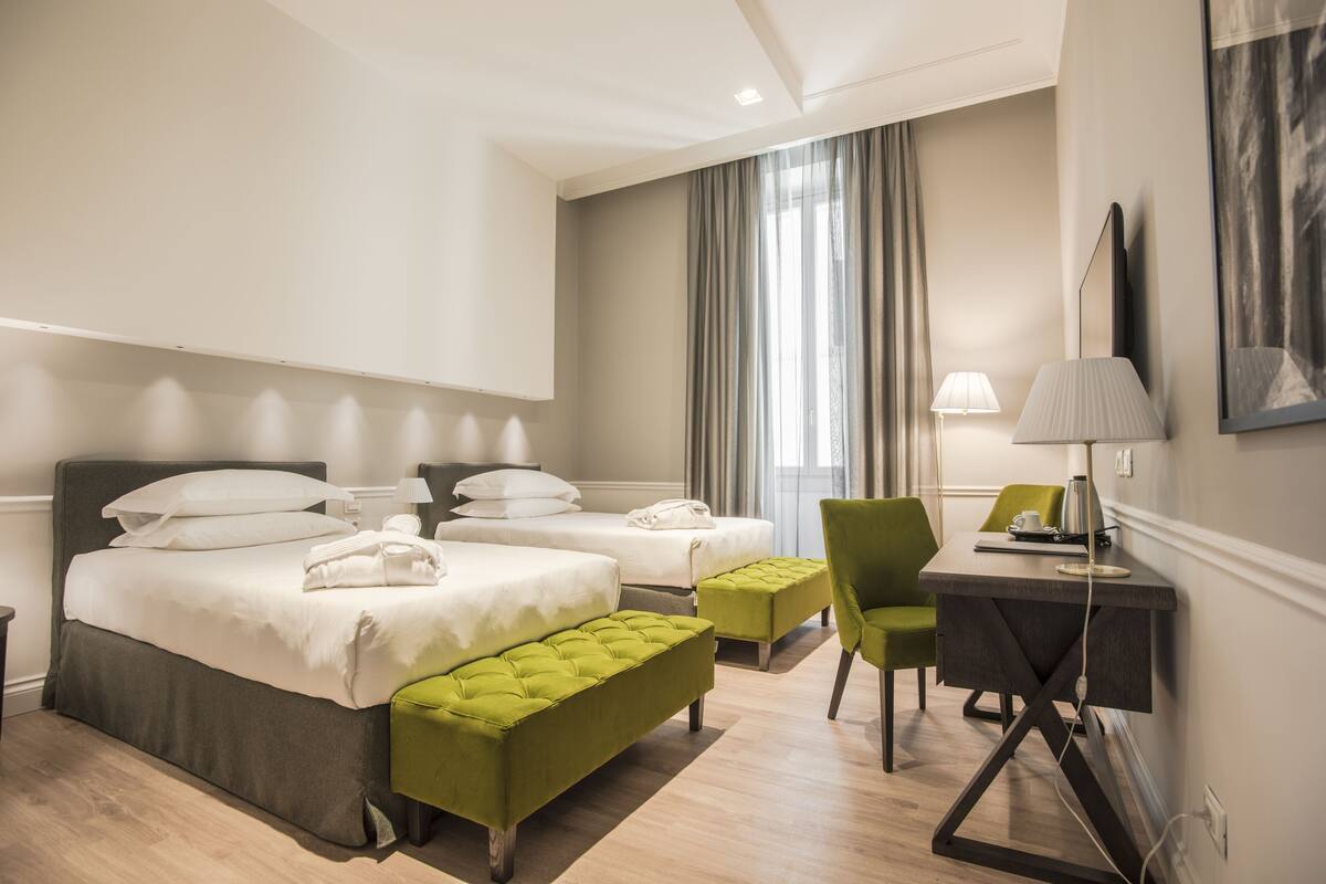 A hotel room with two beds, a desk, a chair, a lamp, and a large window with curtains.