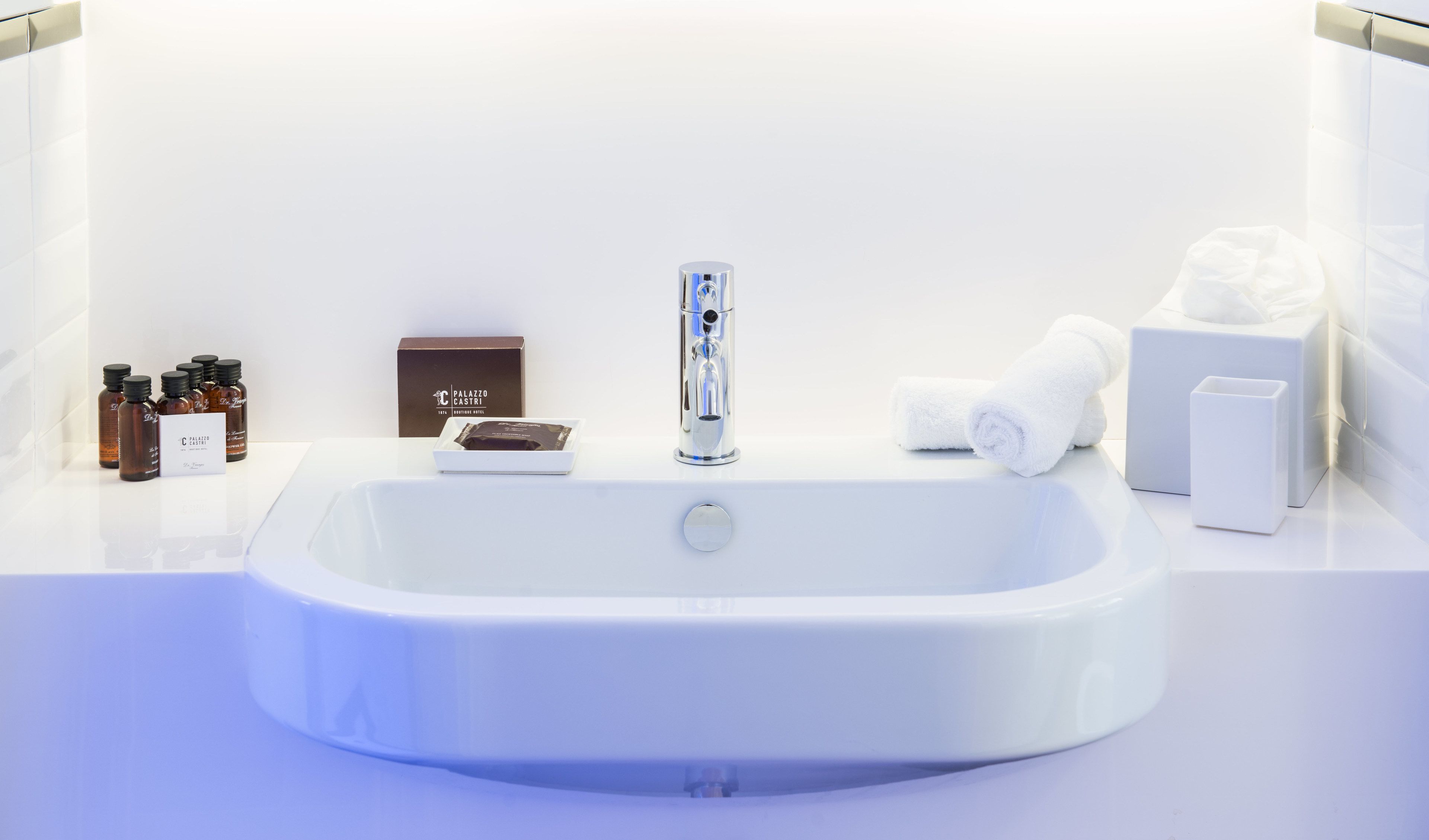 A modern bathroom with a large white sink, a chrome faucet, and a small white cabinet. There are several bottles of toiletries and a box labeled "Palazzo Castri."