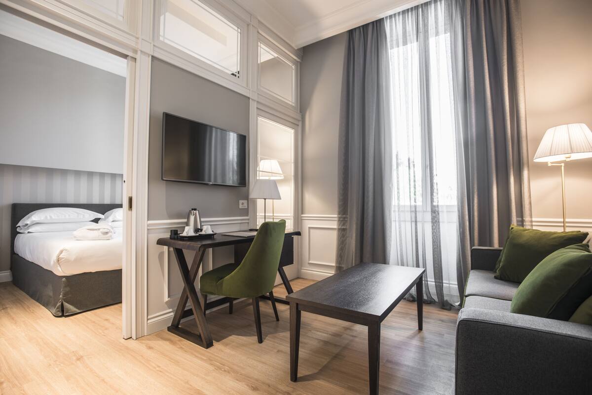 A hotel room with a bed, a desk, a chair, a sofa, a TV, and a lamp.