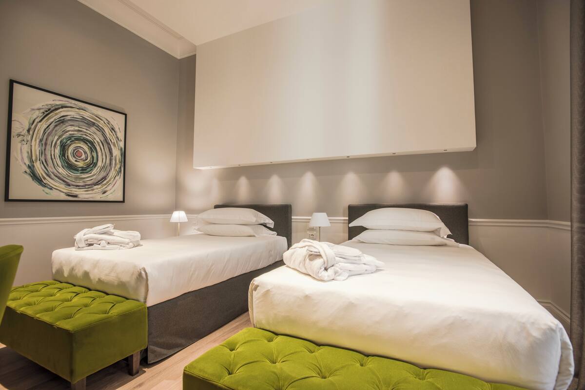 Une chambre d’hôtel avec deux lits, un pouf vert, une œuvre d’art accrochée au mur et une grande fenêtre.