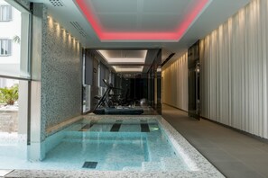 Una piscina interior moderna con pared de cristal, techo con iluminación roja y zona de fitness con equipamiento para ejercicios.