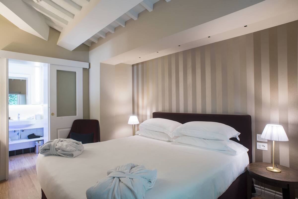 A hotel room with a large bed, two bedside lamps, a robe on the bed, and a door leading to a bathroom.
