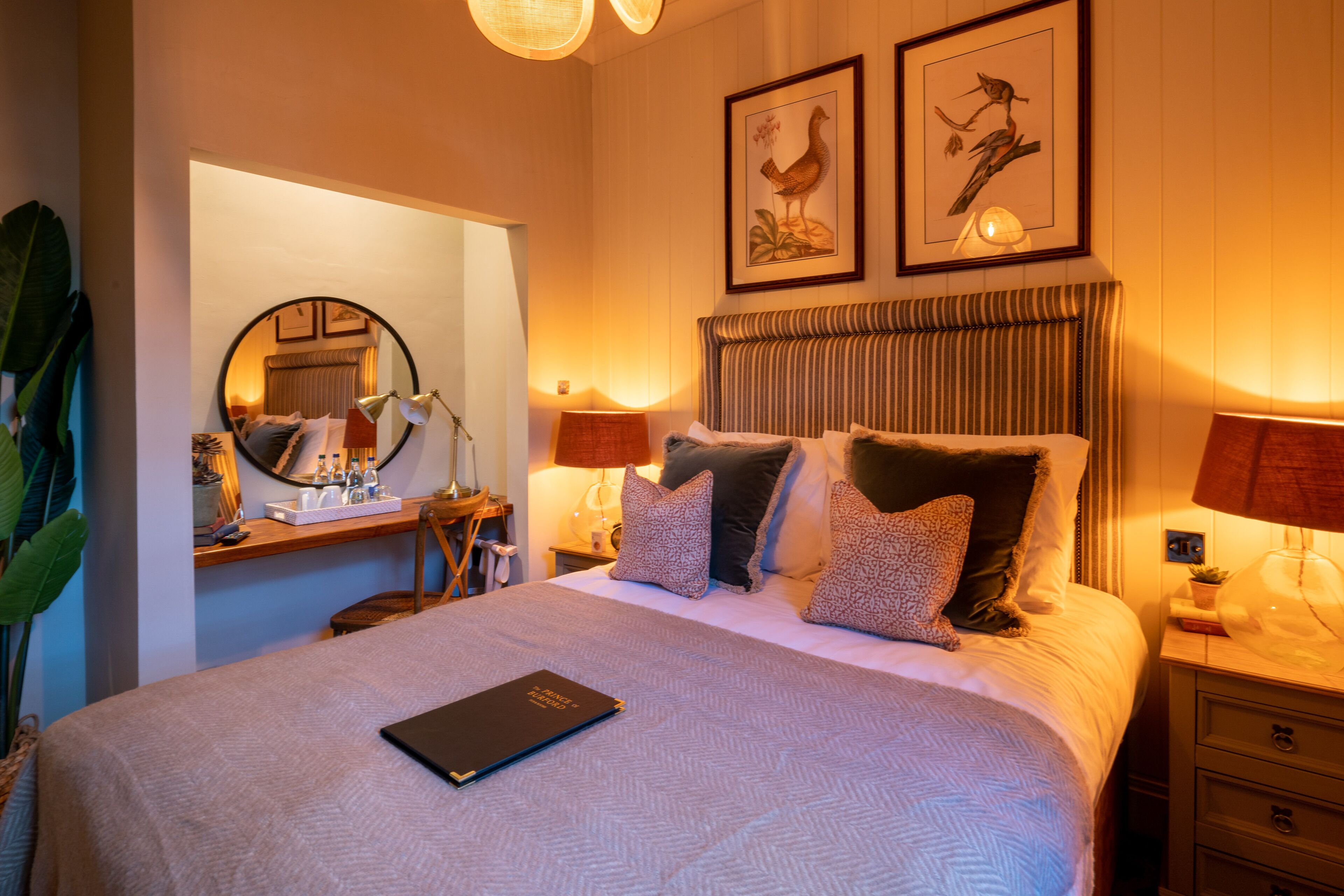 A well-lit bedroom with a large bed, bedside lamps, a bedside table, a mirror, and two framed pictures on the wall.