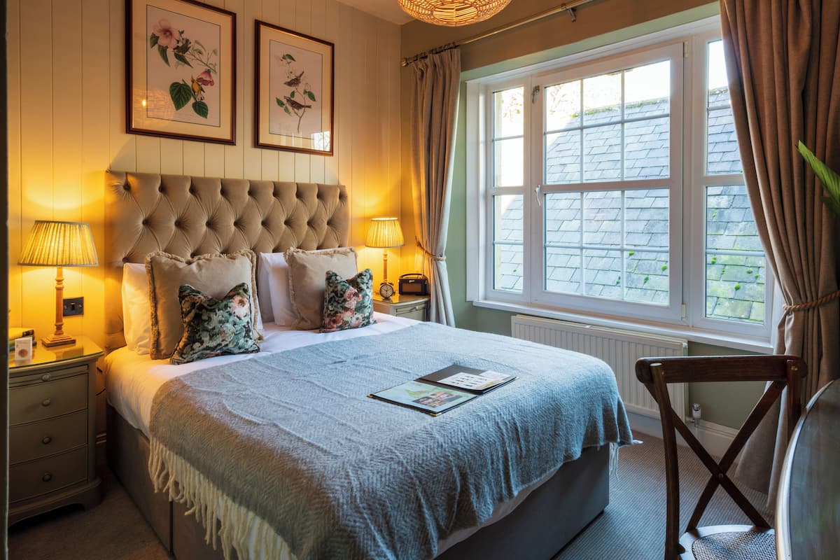 A bedroom with a tufted headboard, a large window with curtains, a bedside table with a lamp, and a chair.