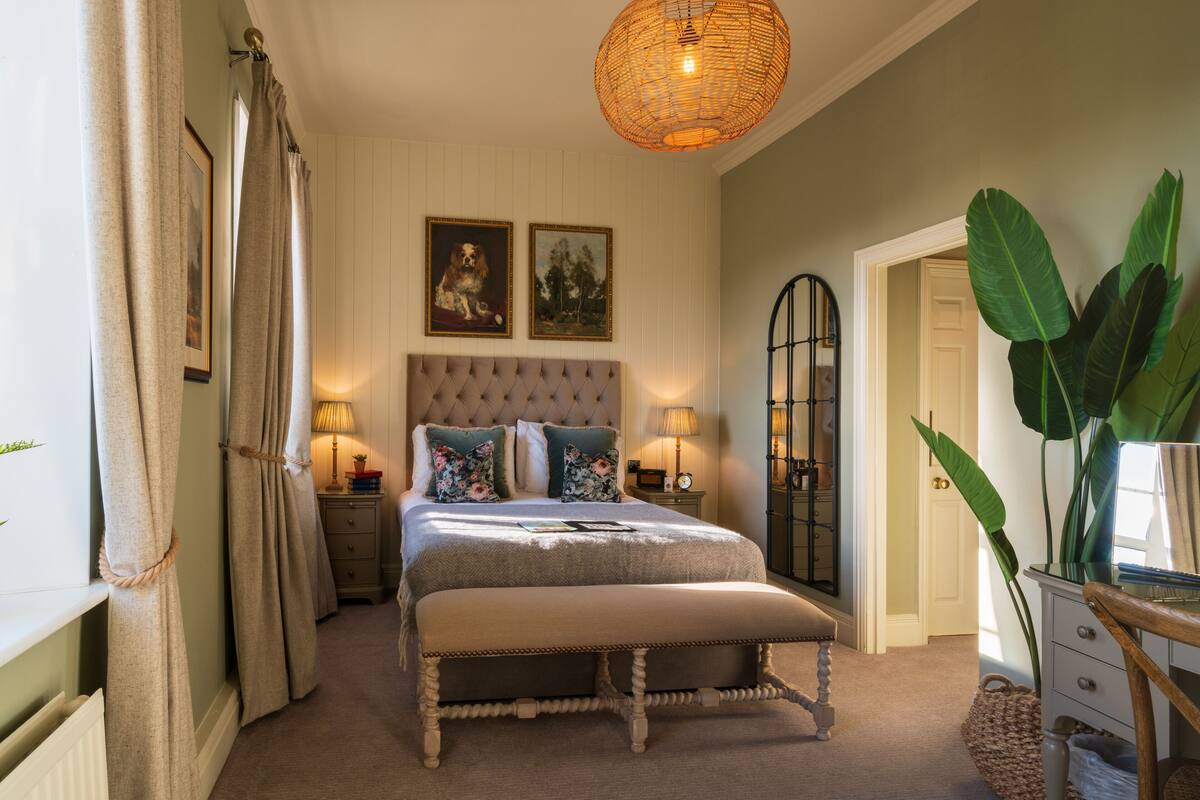 A bedroom with a tufted headboard, a large window with curtains, a bedside table with a lamp, and a large plant.
