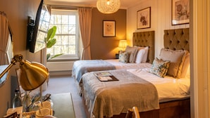 Habitación de hotel con dos camas, un escritorio con lámpara, una ventana con cortinas y cuadros en las paredes.