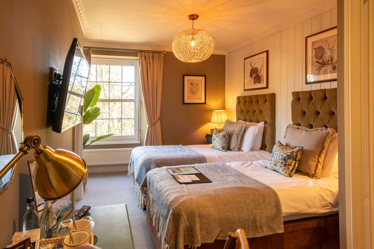 A hotel room with two beds, a desk with a lamp, a window with curtains, and framed artwork on the walls.
