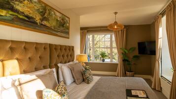 A bedroom with a tufted headboard, a large window with curtains, a television mounted on the wall, and a bed with multiple pillows and a blanket.