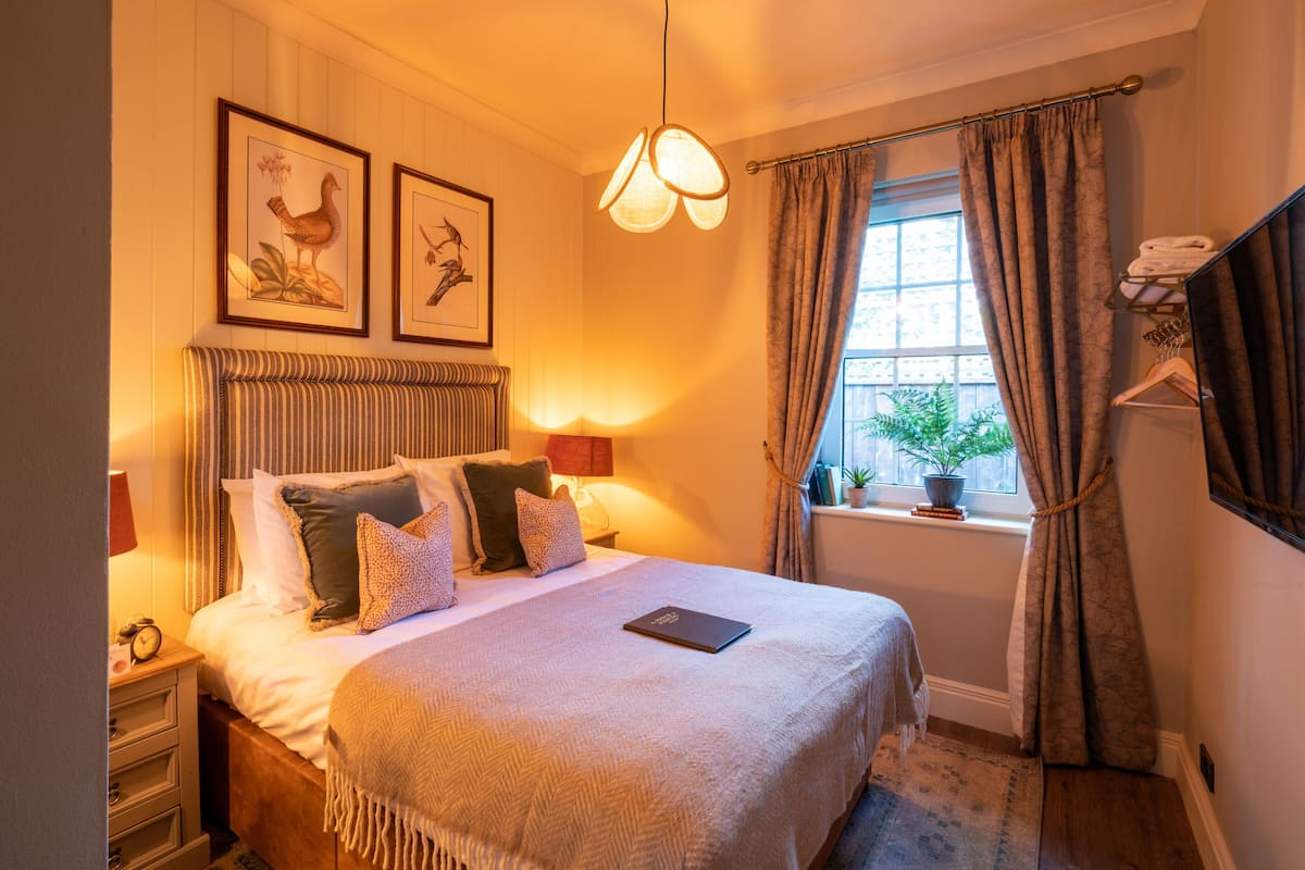 A neatly made bed with a blanket, pillows, and a book on it. There are framed pictures on the wall, a window with curtains, and a shelf with towels.