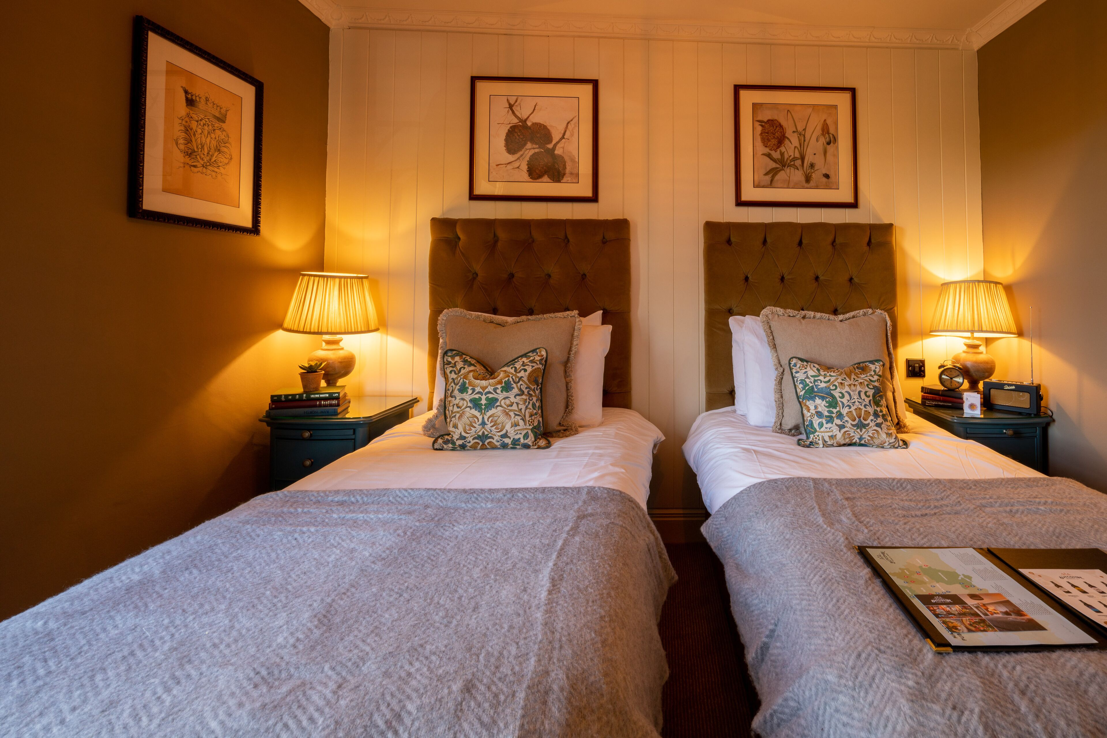 Ein Hotelzimmer mit zwei Betten, jeweils mit einem mit Rautenmuster versehenen Kopfteil, einem Nachttisch mit Lampe und gerahmten botanischen Drucken an der Wand.