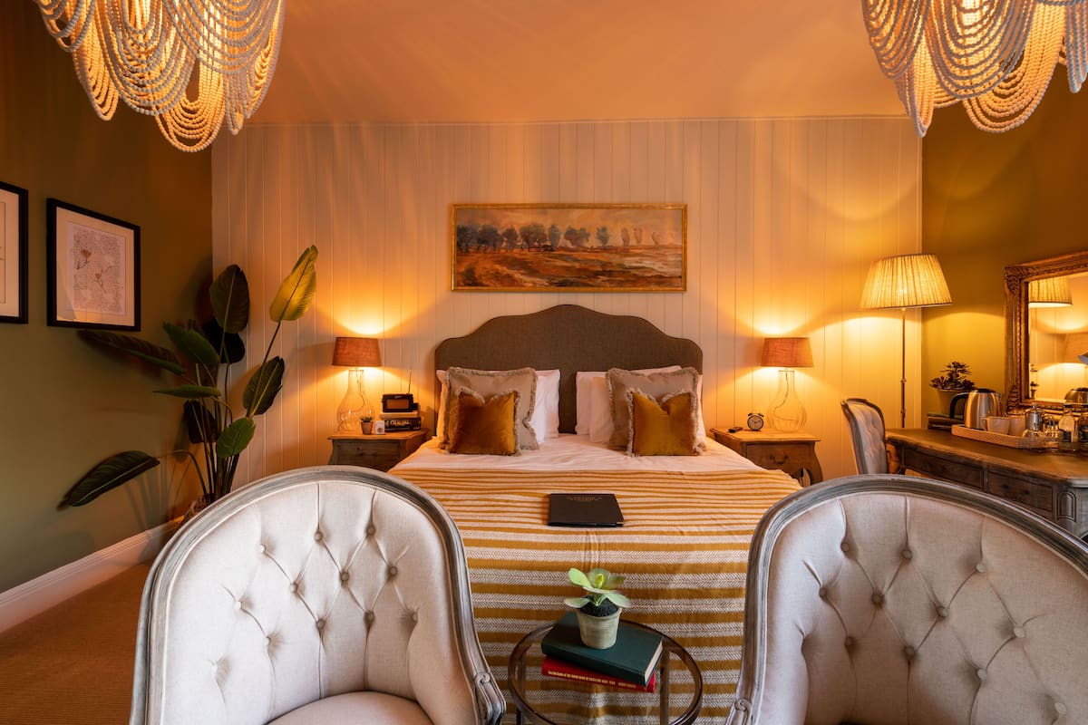 A hotel room with a large bed, two bedside lamps, a painting on the wall, a potted plant, and two armchairs.