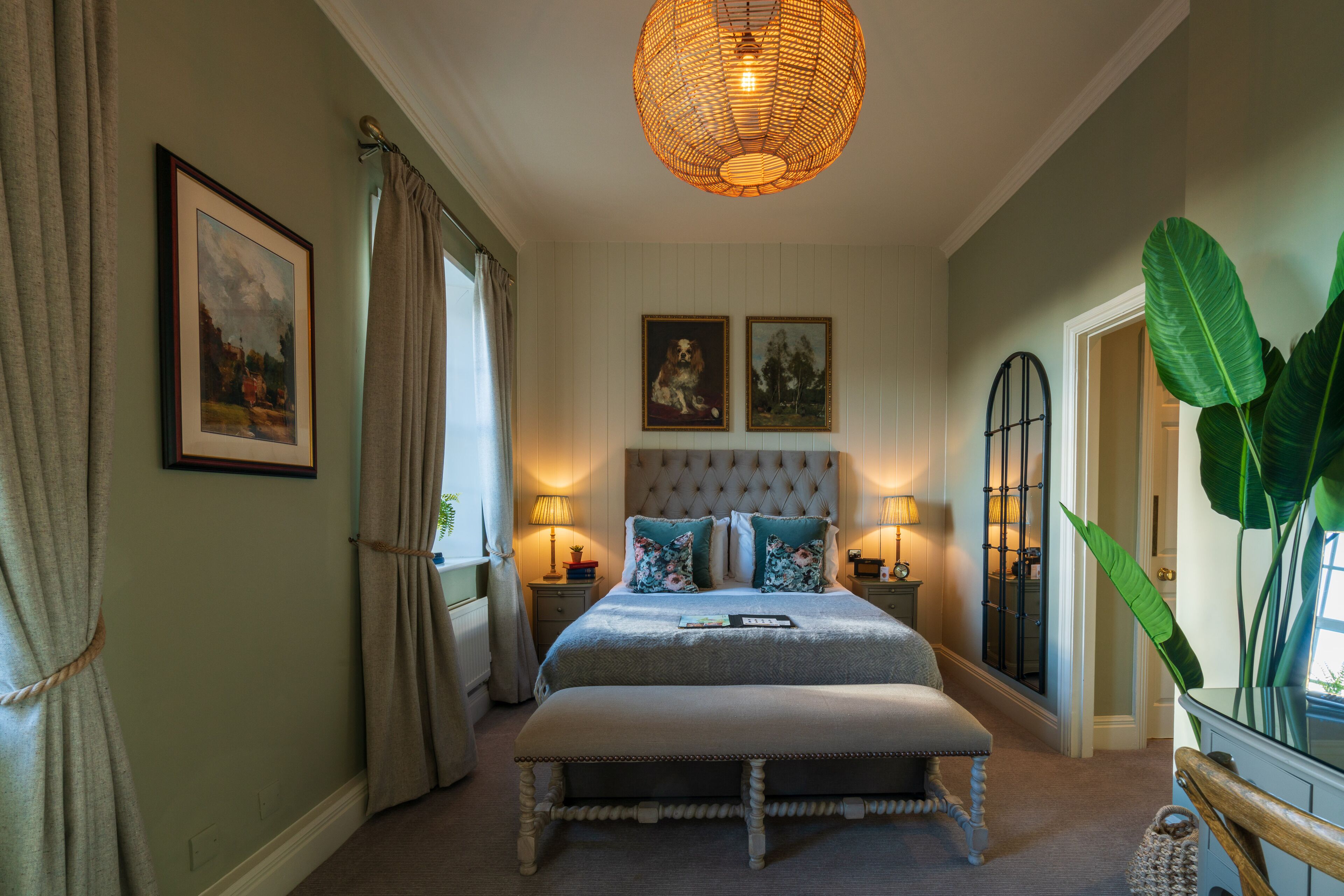 A bedroom with a tufted headboard, a large window with curtains, a bedside table with a lamp, and a large plant.
