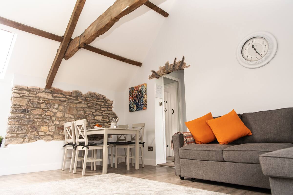 A cozy living room with a stone wall, wooden beams, a dining table with chairs, a grey sofa with orange cushions, and a wall clock.
