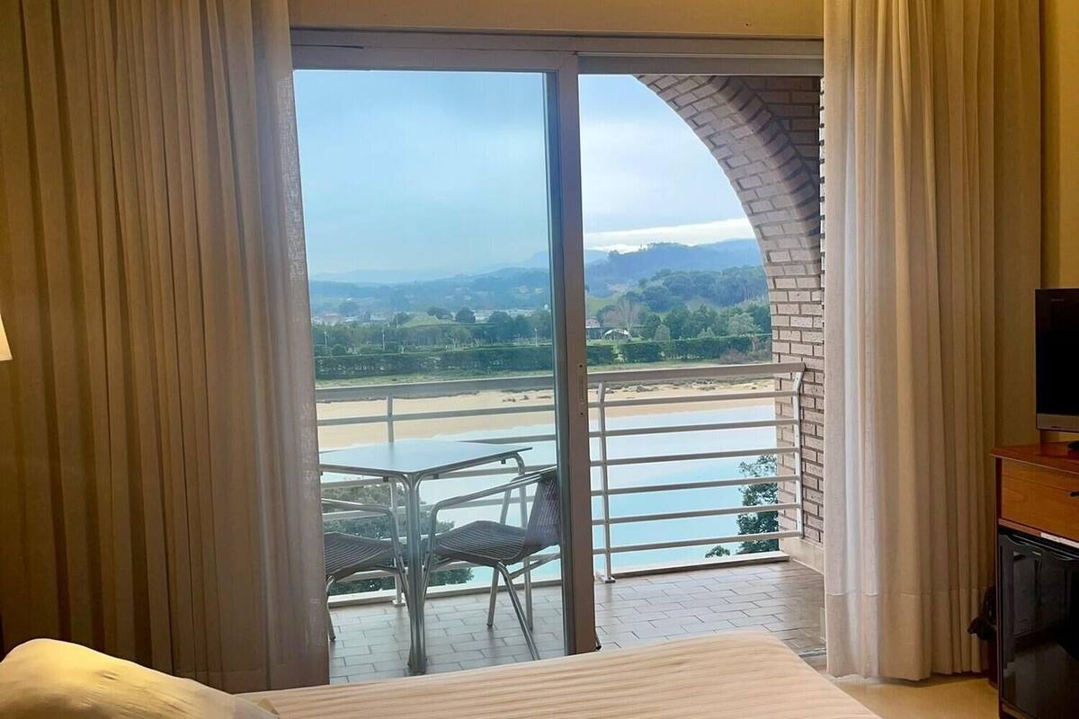 A hotel room with a large bed, a TV, and a balcony with a view of a beach and mountains.