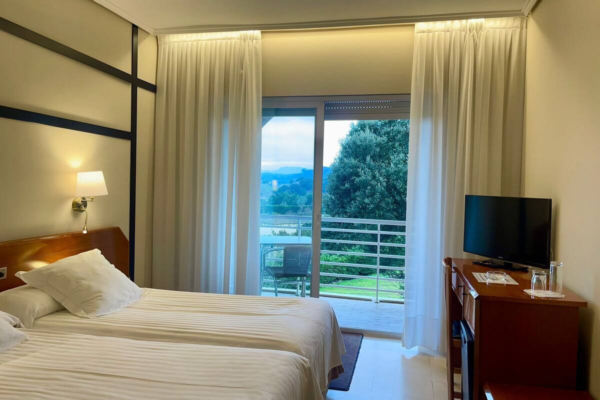 Une chambre d’hôtel avec deux lits, une télévision, un bureau et un balcon donnant sur un espace verdoyant et un immeuble.