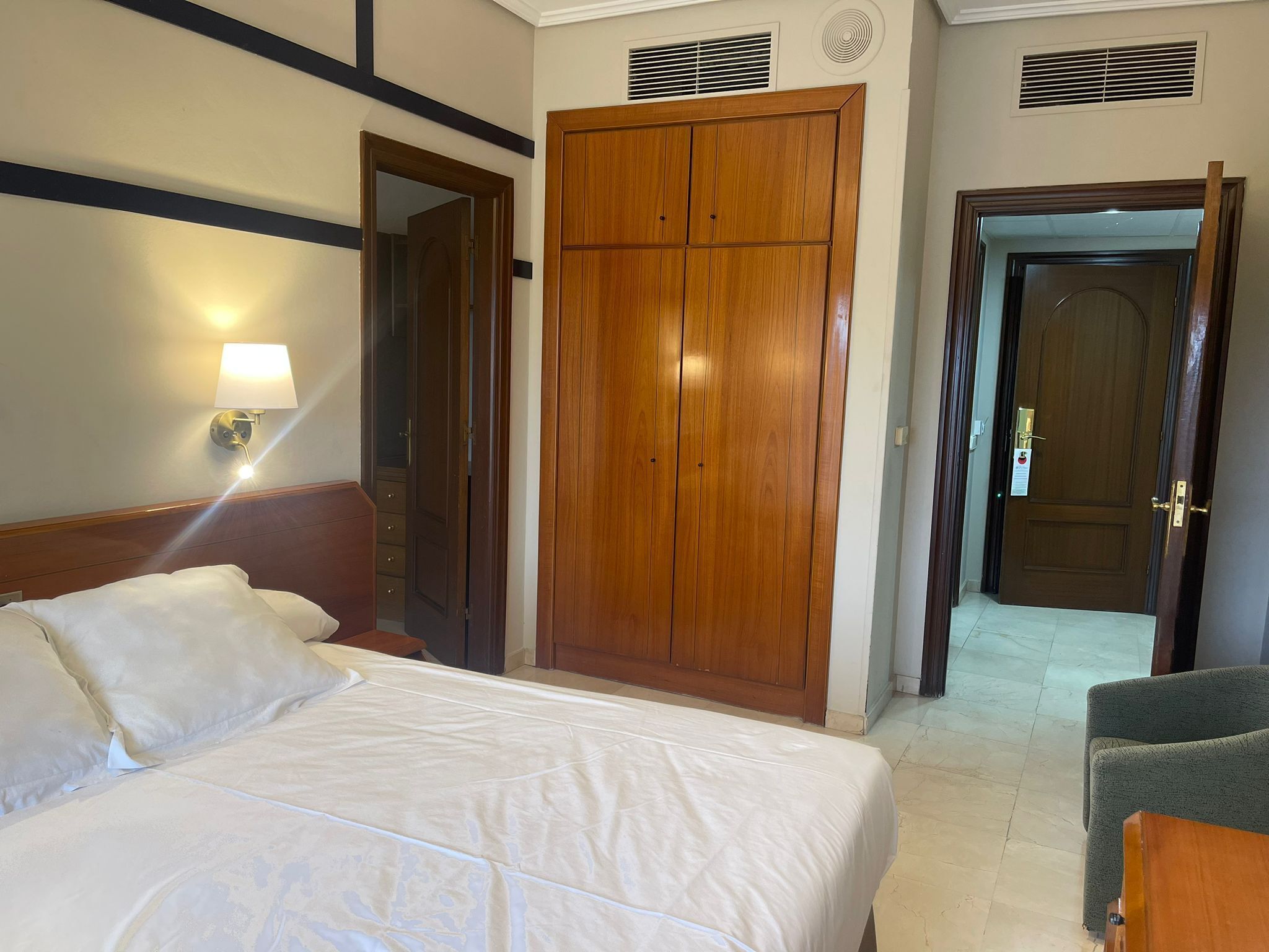 A hotel room with a bed, a wardrobe, a chair, and a wall-mounted lamp.