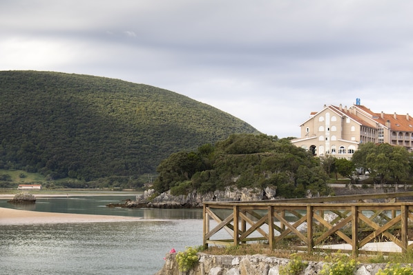 A coastal building with a wooden deck overlooking a body of water and a hill in the background. - Hotel Olimpo (Arnuero)