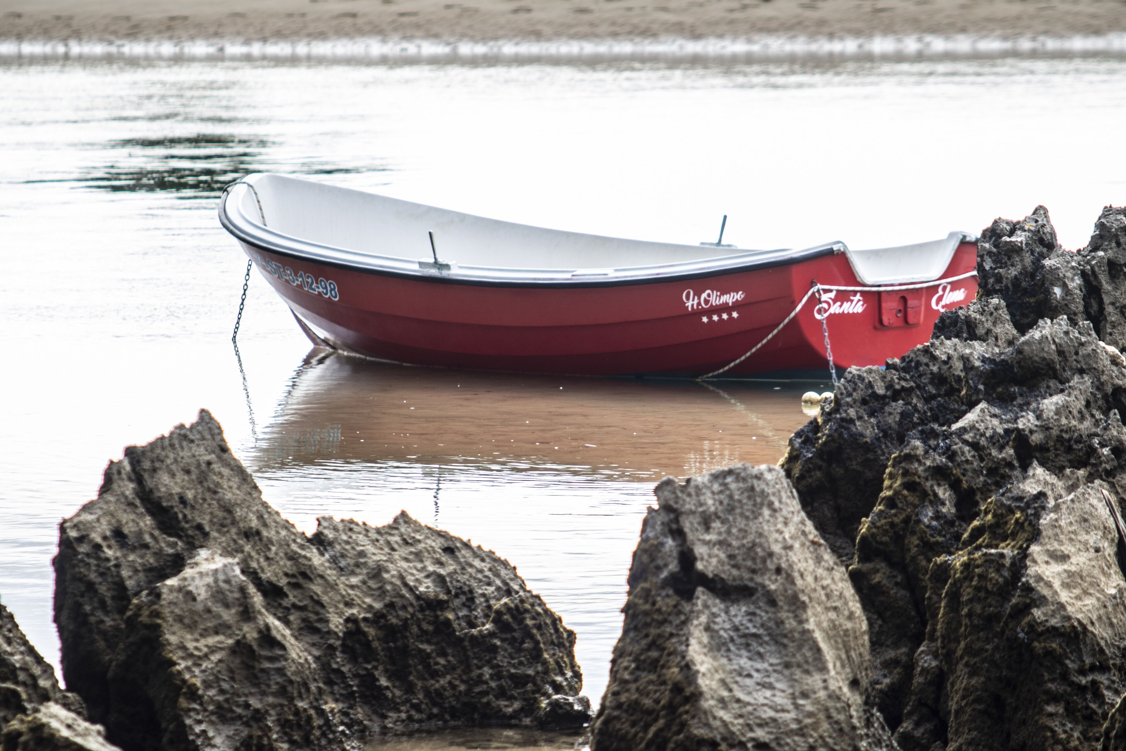 Un barco rojo con el nombre 'Santa' y 'It. Olimpo' escrito, amarrado a una costa rocosa.
