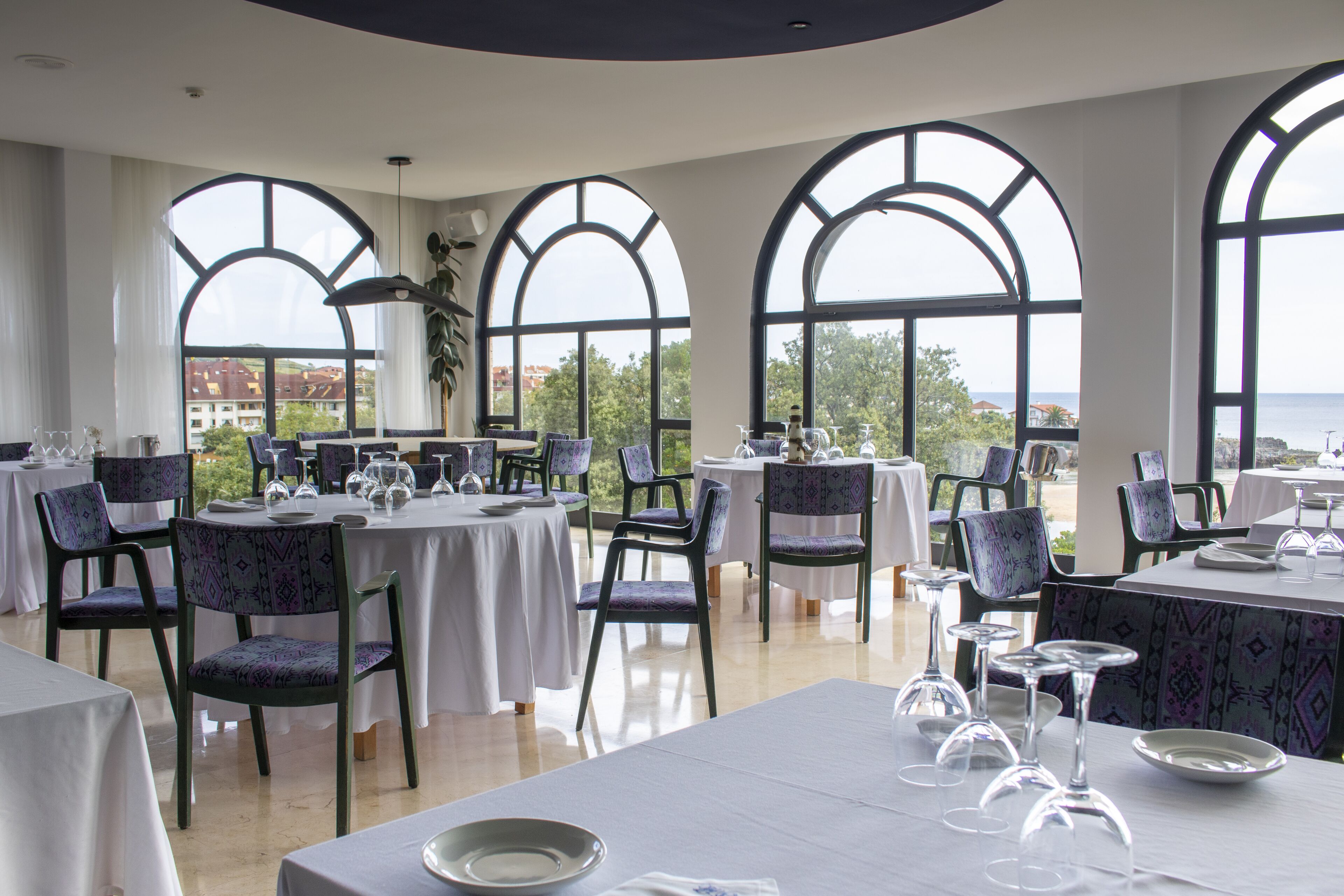 Un restaurante con mesas dispuestas para comer, amplias ventanas de arco y vistas al mar.