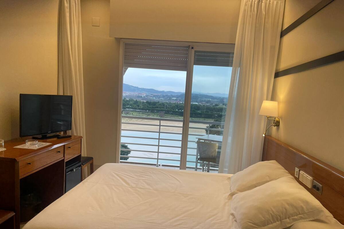 Une chambre d’hôtel avec un lit, un bureau doté d’une télévision, une chaise et une vue sur une rivière et des montagnes, grâce à de grandes fenêtres.