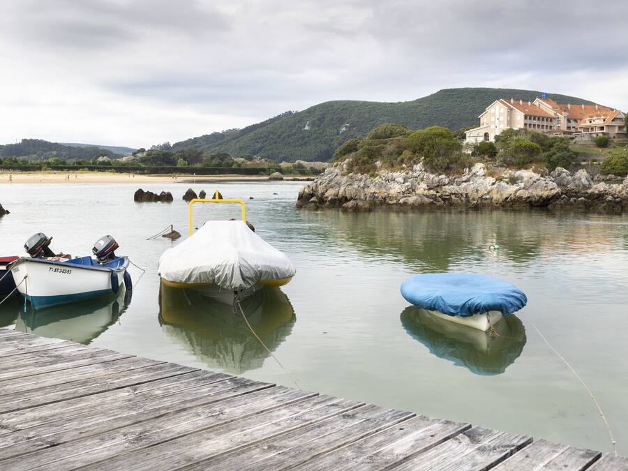 Un muelle de madera con barcos, un lago tranquilo y un edificio grande en una colina al fondo.