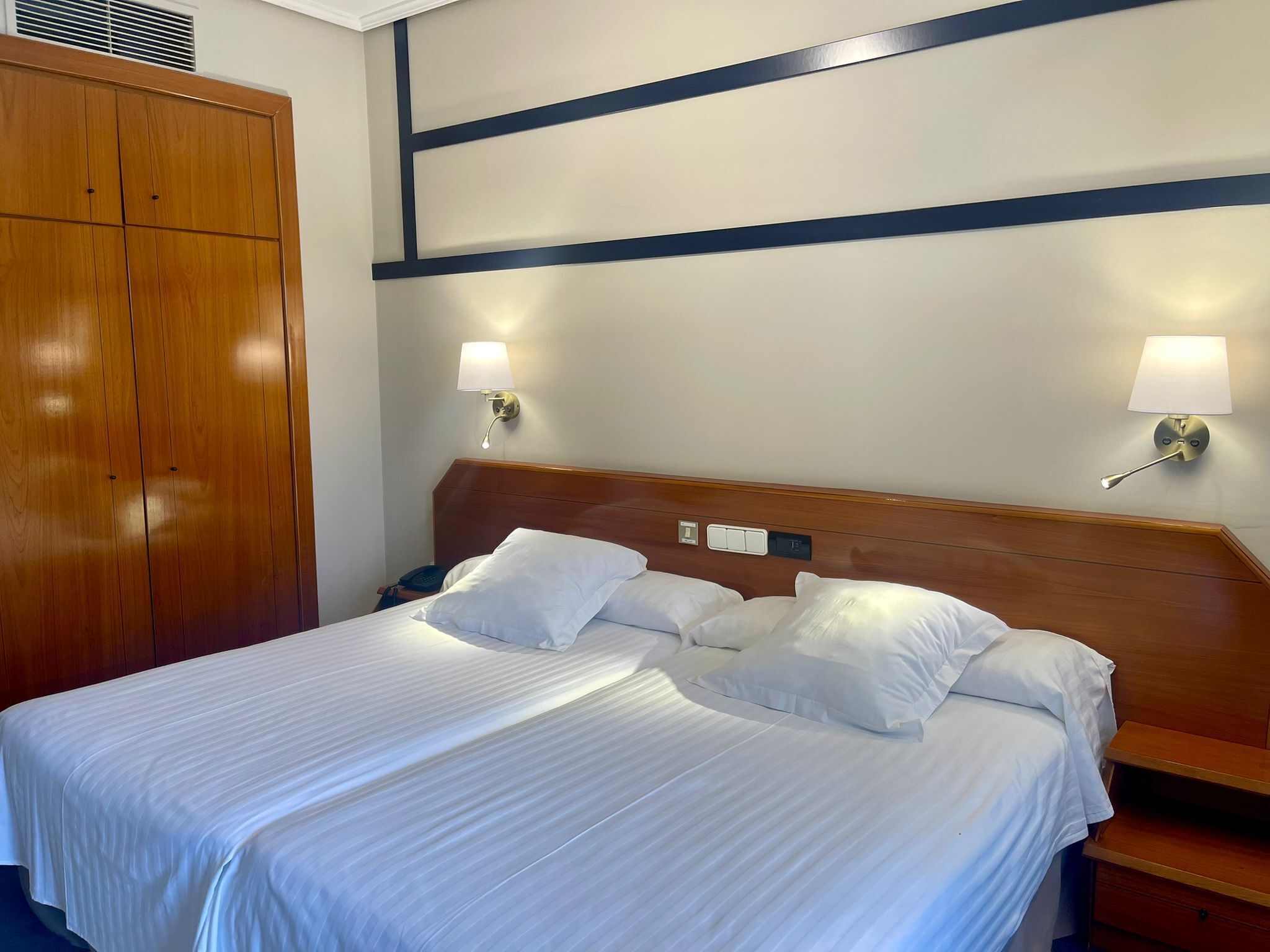 Hotelkamer met een groot bed, twee bedlampen, een houten garderobe en een muur met een geometrisch patroon.
