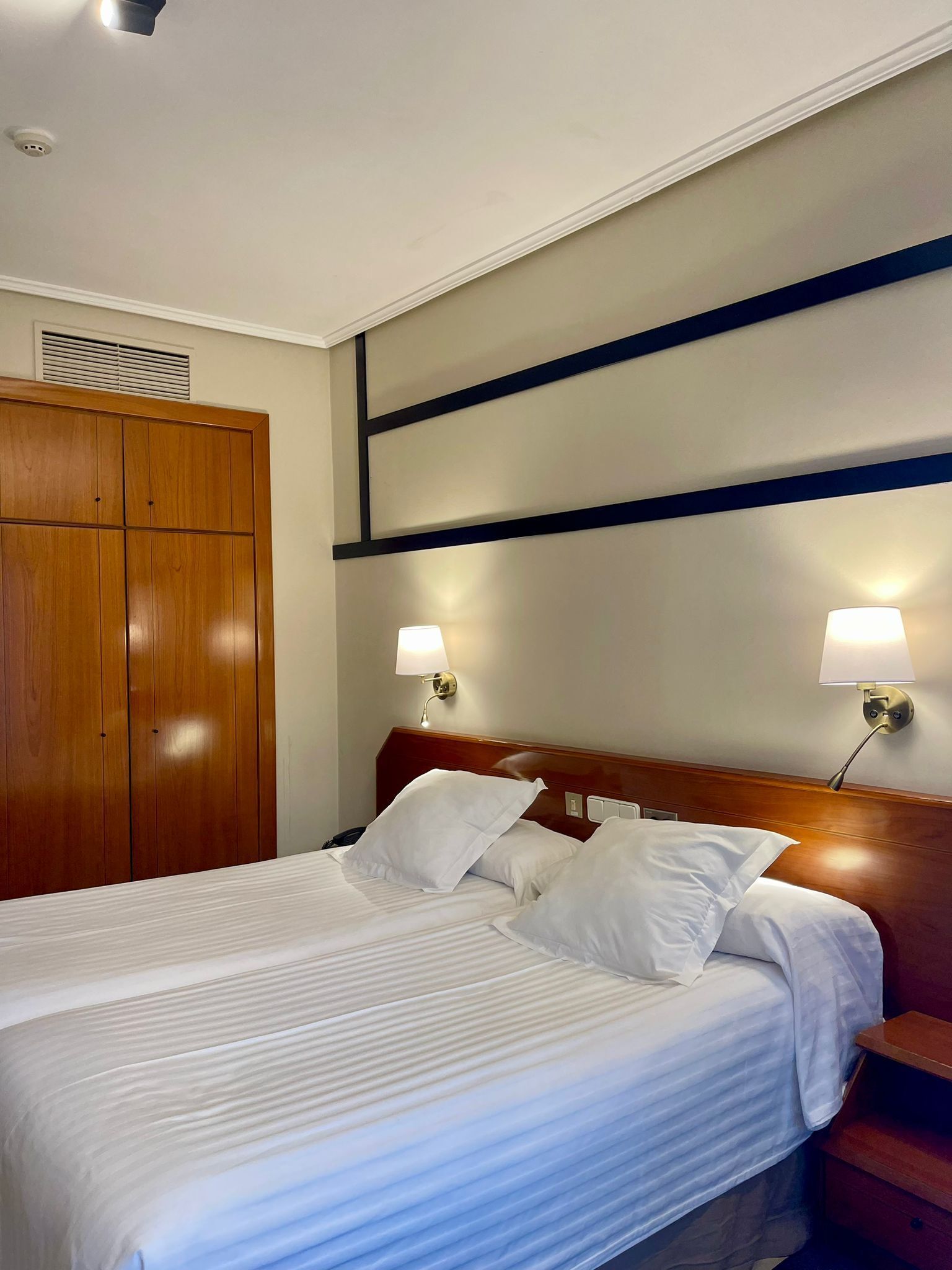 A hotel room with a large bed, wooden wardrobe, and two wall-mounted lamps.