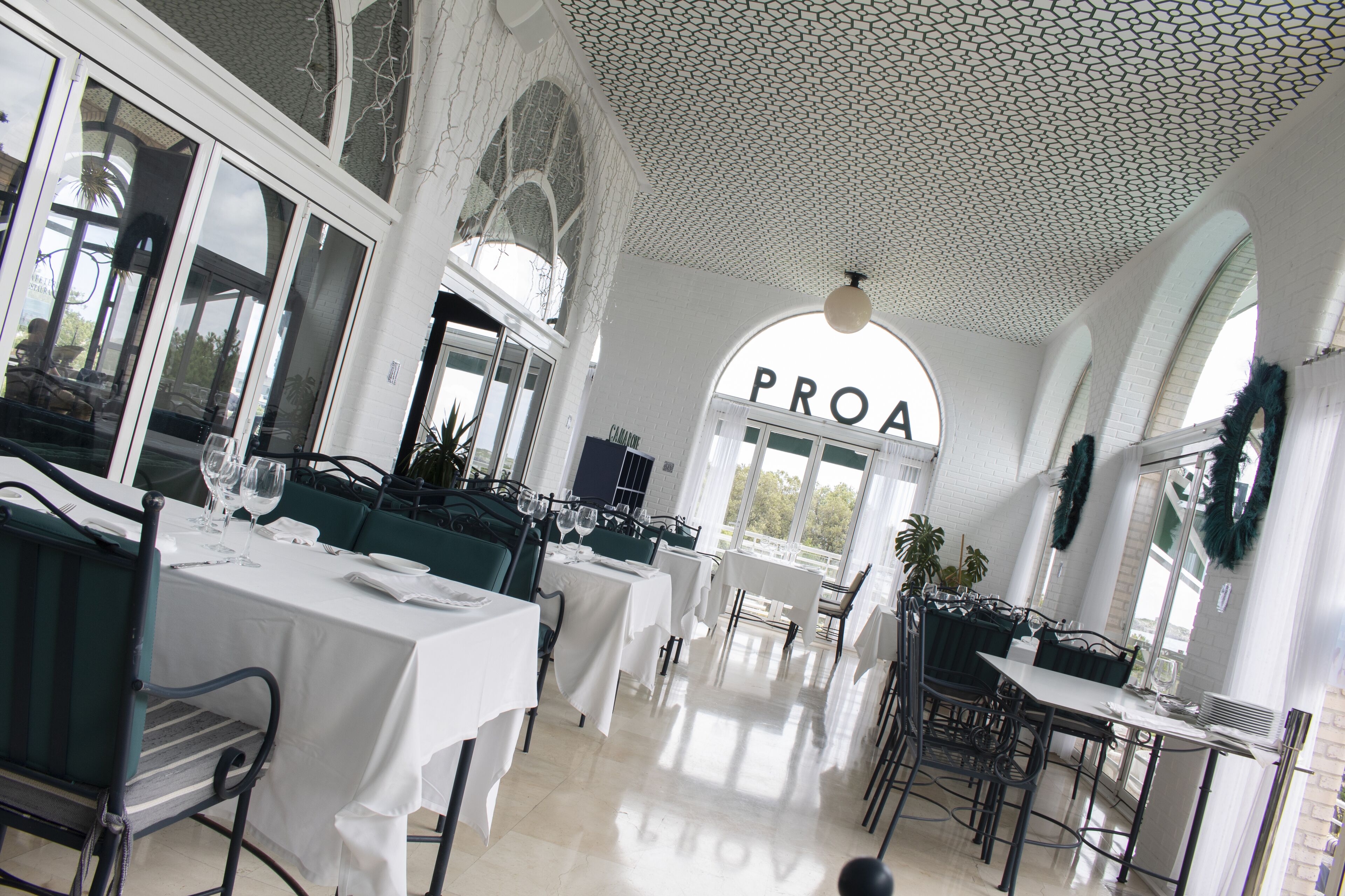 Un restaurante con mesas y sillas blancas, un techo con un diseño geométrico y un amplio ventanal con la palabra "PROA" a la vista.