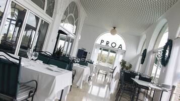 Un restaurante con mesas y sillas blancas, un techo con un diseño geométrico y un amplio ventanal con la palabra "PROA" a la vista.