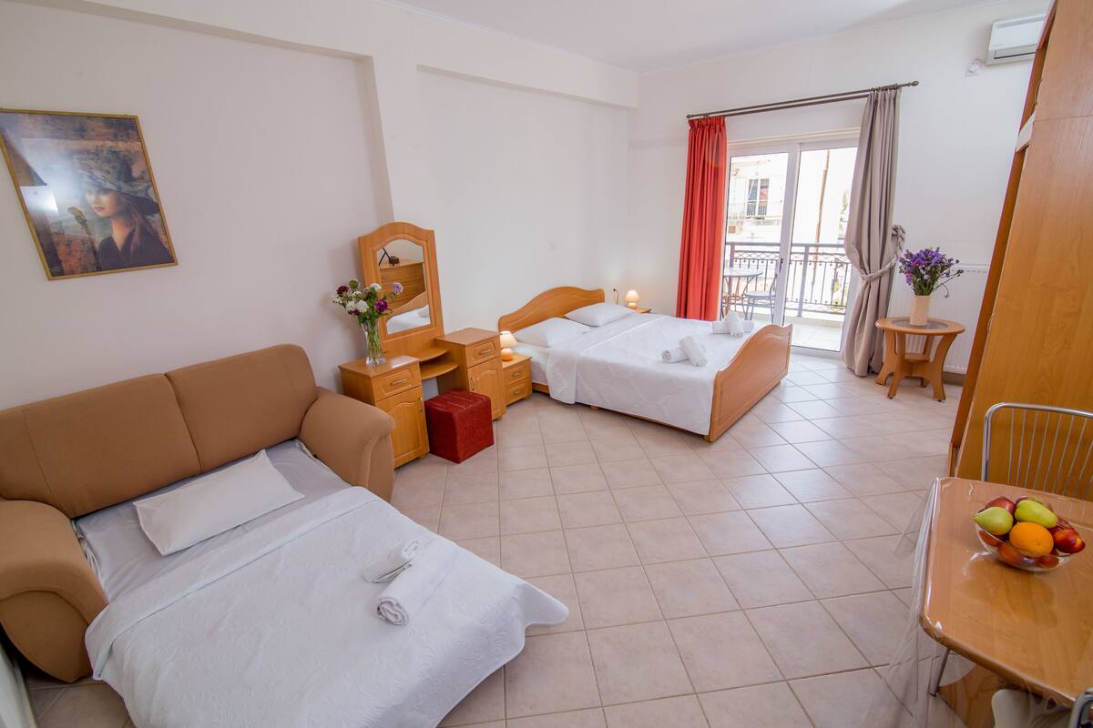 Ein Hotelzimmer mit zwei Betten, einem Schlafsofa, einem kleinen Tisch mit Früchten, einem Spiegel und einem Balkon mit Aussicht.