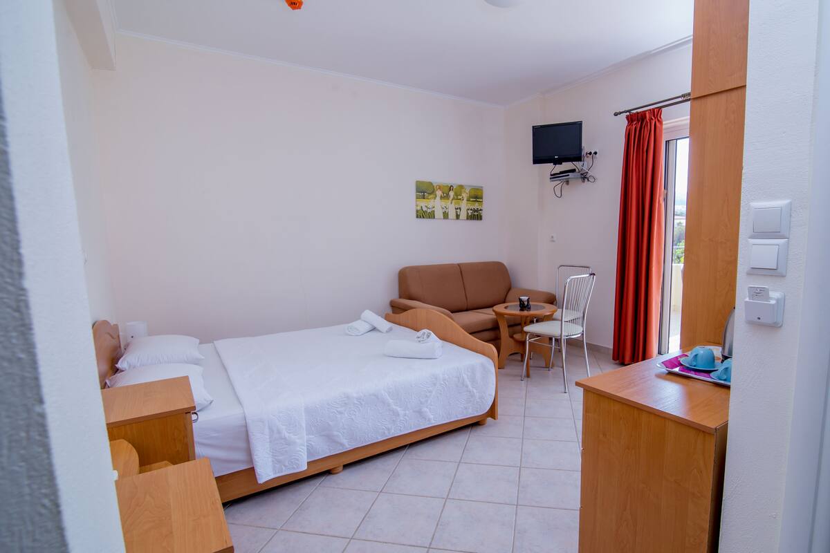 Ein Hotelzimmer mit einem Bett, einem Sofa, einem kleinen Tisch mit Stuhl, einem an der Wand befestigten Fernseher und einem Fenster mit Vorhängen.