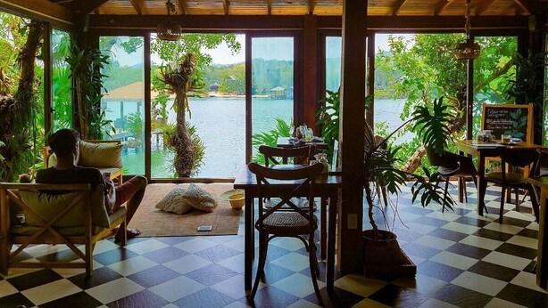 A room with a checkered floor, wooden furniture, a table with chairs, and large windows offering a view of a body of water and greenery. - Hotel Posada Las Orquideas (Roatan)